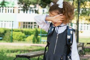 Girl with a backpack near the school after classes, unhappy, tired and headache. The child is crying, holding his head in his hands. Academic failures, poor grades, overwork photo