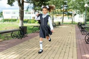 alegre gracioso niña con sin dientes sonrisa en colegio uniforme con blanco arcos corriendo en colegio patio trasero. espalda a escuela, septiembre 1. contento alumno con mochila. primario educación, elemental clase. movimiento foto