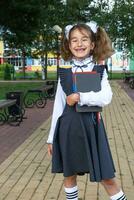 Girl with backpack, school uniform with white bows and stack of books near school. Back to school, happy pupil, heavy textbooks. Education, primary school classes, September 1 photo