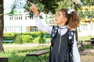 Cheerful funny girl in a school uniform with white bows in school yard points with his finger. Back to school, September 1. Happy pupil with a backpack. Primary education, elementary class. photo