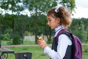 Girl with a backpack eating pie near school. A quick snack with a bun, unhealthy food, lunch from school. Back to school. Education, primary school classes, September 1 photo