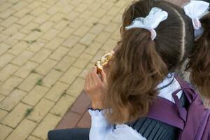 niña con un mochila comiendo tarta cerca escuela. un rápido bocadillo con un bollo, insalubre alimento, almuerzo desde escuela. espalda a escuela. educación, primario colegio clases, septiembre 1 foto