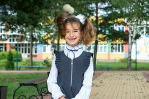 Cheerful funny girl with a toothless smile in a school uniform with white bows in school yard. Back to school, September 1. A happy pupil. Primary education, elementary class. Portrait of a student photo