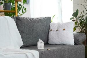 Key to home with keychain and and miniature of house on gray sofa in white interior with potted plants. Design, project, moving to new house, mortgage, rent and purchase real estate photo
