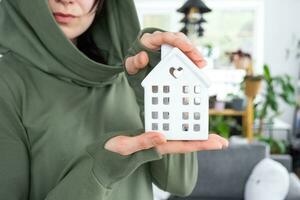 A happy woman in her house holds in her hands a miniature figure of a house and key in the interior. Dream house project, real estate purchase, insurance, mortgage, rent, reservation photo