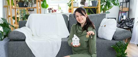 A happy woman in her house holds in her hands a miniature figure of a house and key in the interior. Dream house project, real estate purchase, insurance, mortgage, rent, reservation photo