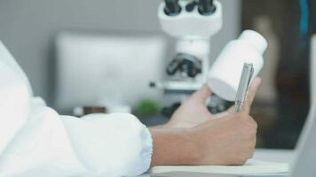 Medical worker in lab coat and sterile mask, doing a microscope analysis while her colleague are working behind video