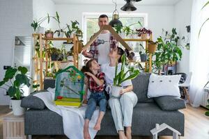 traditional family with a child and a parrot pet in a cage are enjoying a new home, sitting on the sofa and holding the roof. Mortgage, insurance, buying and moving into your own home photo