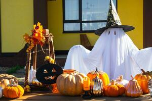 Child in bed sheet with slits like ghost in witch's hat and Halloween decor on the porch of the house outside in the yard of pumpkin, lantern, garlands, ack lantern. party, autumn mood photo