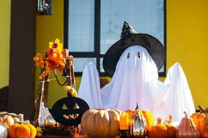 Child in bed sheet with slits like ghost in witch's hat and Halloween decor on the porch of the house outside in the yard of pumpkin, lantern, garlands, ack lantern. party, autumn mood photo