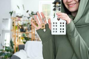 A happy woman in her house holds in her hands a miniature figure of a house and key in the interior. Dream house project, real estate purchase, insurance, mortgage, rent, reservation photo