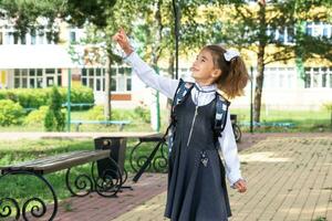 Cheerful funny girl in a school uniform with white bows in school yard points with his finger. Back to school, September 1. Happy pupil with a backpack. Primary education, elementary class. photo