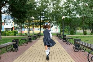 Cheerful funny girl with toothless smile in school uniform with white bows running in school yard. Back to school, September 1. Happy pupil with backpack. Primary education, elementary class. motion photo