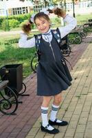 Cheerful funny girl with a toothless smile in a school uniform with white bows in school yard. Back to school, September 1. Happy pupil with a backpack. Primary education, elementary class. photo