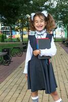 Girl with backpack, school uniform with white bows and stack of books near school. Back to school, happy pupil, heavy textbooks. Education, primary school classes, September 1 photo