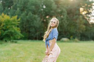 Young Caucasian woman or girl in a summer dress and a denim shirt is walking on the grass, holding shoes in her hands, relaxing outside the city on a weekend in summer photo