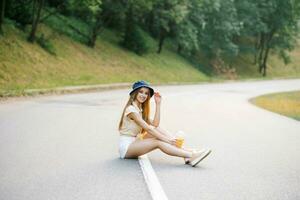 A happy girl in white shorts and a yellow blouse holds a glass of juice with a straw in her hands and sits on the road near the forest photo