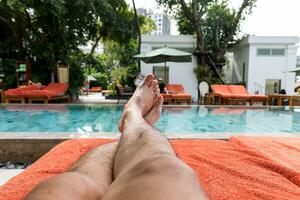 broncearse por el hotel turista hotel nadando piscina, mans piernas acostado abajo en un tumbona mirando terminado el agua foto
