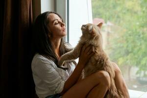 linda mujer sentado en el antepecho con un rojo gato y mirando fuera el ventana foto