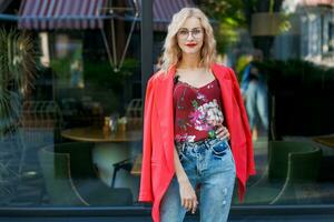 beautiful blonde woman in glasses dressed in red jacket and blue jeans posing on street in the city photo