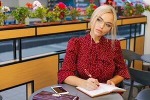 bonito caucásico mujer escribe en cuaderno gasto hora al aire libre en cafetería. rubia foto