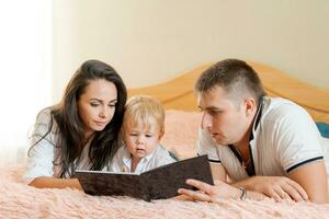 contento familia acostado en el cama leyendo un libro, mamá papá y pequeño hijo foto