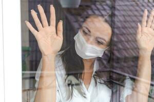 woman with a protective mask looks out the window with longing photo