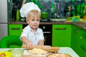 contento pequeño chico preparando masa en cocina a mesa. allí son masa productos en mesa, vestido como cocinero foto