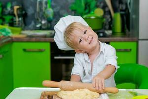 contento pequeño chico preparando masa en cocina a mesa. allí son masa productos en mesa, vestido como cocinero foto