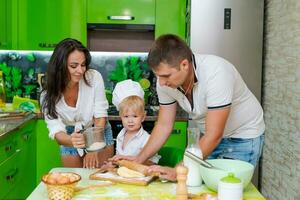 contento familia mamá y pequeño hijo y papá son preparando masa en cocina a mesa. productos para masa son en mesa foto