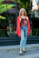 beautiful blonde woman in glasses dressed in red jacket and blue jeans posing on street in the city photo