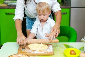 happy little boy preparing dough in kitchen at table. there are dough products on table, dressed as chef photo