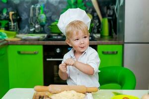 happy little boy preparing dough in kitchen at table. there are dough products on table, dressed as chef photo
