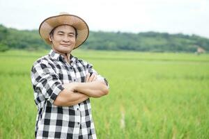 Asian man farmer , wears hat, plaid shirt, stands at paddy field, crossed arms on chest with confident. Concept, agriculture occupation. Thai farmer lifestyle. Visit and take care of crops photo