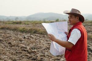 Asian man explorer wears hat, red vest shirt, holds map to explore land boundary. Concept, land planning, exploring property. Geodetic survey area. Treasure hunting on land. Adventure lifestyle. photo