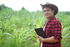 Handsome Asian man farmer is in garden, wears hat, red plaid shirt, holds  smart tablet. Feel confident, proud in crops. Concept, Agriculture occupation, inspect, survey and research. Copy space. photo