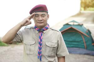 andsome Asian man wear boy scout uniform red cap blue and pink striped scarf, make hand sign symbol at outdoor campingsite.Concept, educational career with uniform in school, Thailand. Go camping photo