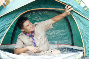 Handsome Asian man wears boy scout uniform sits in camping tent. Concept, accommodation for temporary outdoor sleeping as scout hiking activity. Scout go camping. photo
