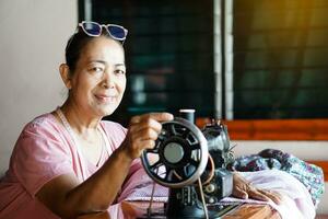 Happy senior Asian woman tailor is sewing cloth by vintage sewing machine at home. Concept, Senior  lifestyle, spend  free time to repair or design clothes. Hobby, not lonely. photo