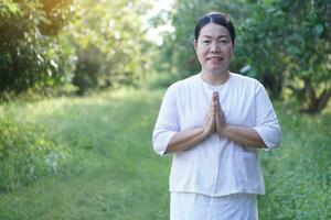 asiático mujer budista usa blanco traje, poner palmas juntos para Orando, soportes a tranquilo bosque. concepto, cultura de fe. religioso actividad. paz de mente gestión. foto