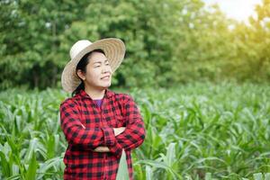 Asian farmer wears hat, red plaid shirt, cross arms on chest, stands at garden. feels confident. Concept, Agriculture occupation. Thai farmer. Working with nature. Organic farming. photo