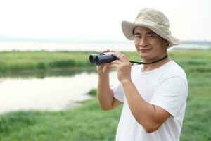 Handsome Asian man ecologist is surveying nature at the lake, hold binoculars. Concept, nature exploration. Ecology study. Pastime activity, lifestyle. Explore environment photo