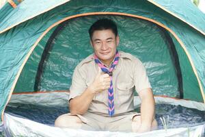 Handsome Asian man wears boy scout uniform sits in camping tent. Concept, accommodation for temporary outdoor sleeping as scout hiking activity. Scout go camping. photo