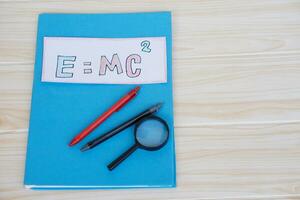 Paper card of formula E MC2 on colorful books with pens, water color tray, crayons and magnifying glass. Concept, Einstein formula. Mass Energy Equivalence. Education, physics subject. photo