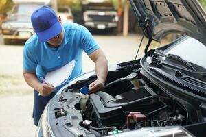 Closeup man mechanic inspecting car engine. holds paper, checking and analyzing car engine under the hood. Concept, Outdoor car inspection service. Claim for accident insurance. photo