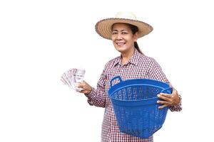 Happy Asian senior woman farmer wears hat, holds blue basket and Thai banknote money, isolated on white background. Concept, Farmer happy to get profit, income from doing agriculture. photo