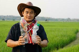 Asian farmer picking up used plastic bottle garbage at green paddy field that cause the problems and pollution for farming. Green Environmental agriculture concept. Reduce, reuse, recycle garbage. photo