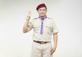 Handsome Asian man wear boy scout uniform, red cap, blue and pink striped scarf, make hand sign symbol of scout for oath. Concept, educational career with uniform in school, Thailand. photo