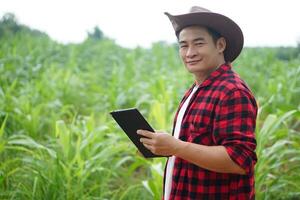 Handsome Asian man farmer is in garden, wears hat, red plaid shirt, holds  smart tablet. Feel confident.Concept, Agriculture occupation, inspect, survey and research about agricultural crops. photo