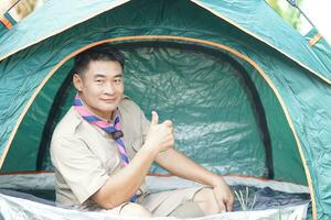 Handsome Asian man wears boy scout uniform sits in camping tent. Concept, accommodation for temporary outdoor sleeping as scout hiking activity. Scout go camping. photo